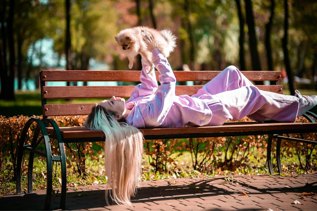 lady laying down on a park bench holding up a pomeranian above her head